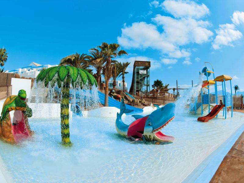 Aire de jeux aquatique colorée avec palmiers, toboggans et arroseurs sous un ciel bleu.