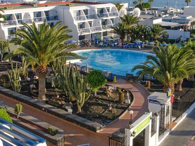 Hotelanlage mit großem Pool, Palmen, weißem Gebäude und Blick auf das blaue Meer im Hintergrund.