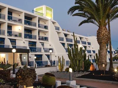 Modern hotel met terras, palmbomen en vetplanten op de voorgrond onder een heldere hemel.