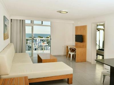 Helles Wohnzimmer mit weißem Sofa, Holzmöbeln, Flachbildschirm und Balkon mit Meeresblick.