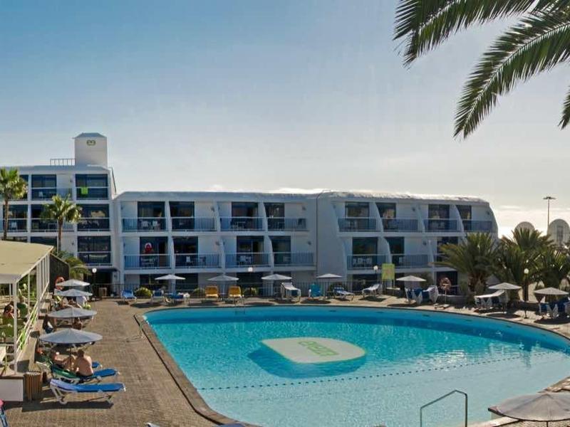 Großer Pool mit blauem Wasser, Sonnenliegen, Palmen und modernem Hotelgebäude im Hintergrund.