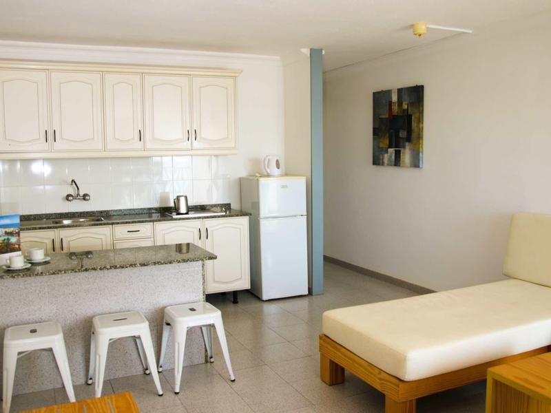 Kleine Küche mit weißer Einrichtung, weißem Kühlschrank, Barhockern und beige Couch im Wohnzimmer.