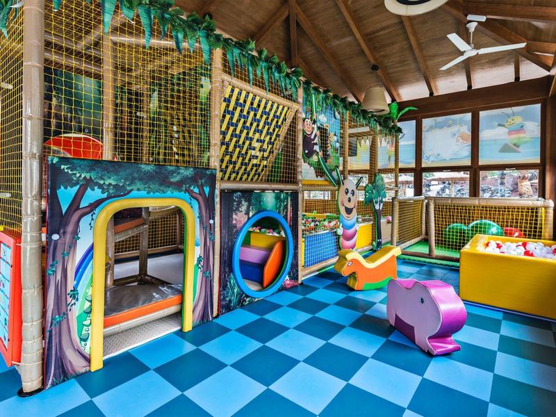 Area giochi interna colorata con struttura per arrampicata, vasca di palline e giocattoli su pavimento blu.