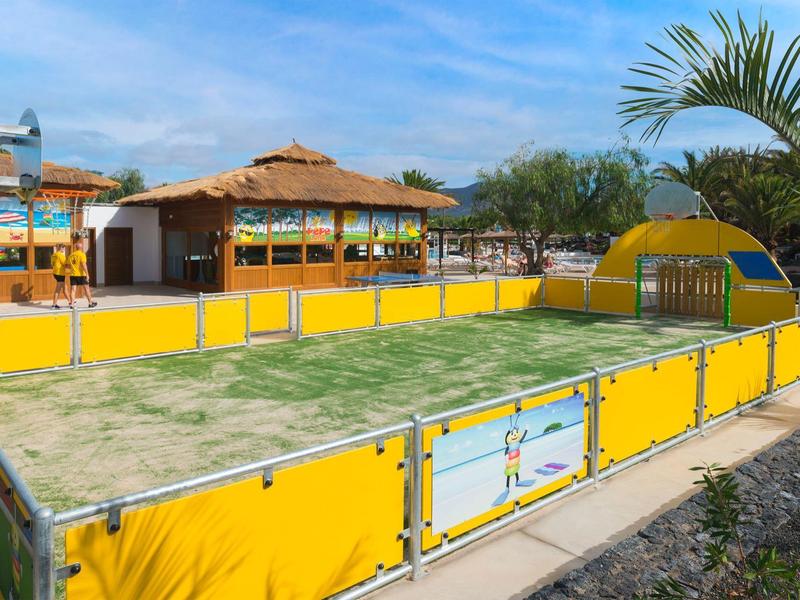 Piccolo campo recintato con bordo giallo vicino a palme e edifici.