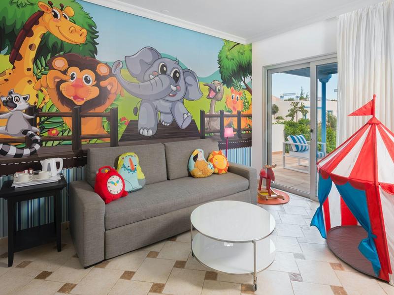 Camera per bambini con murale di animali, divano grigio, tenda da circo colorata e balcone con vista piscina.