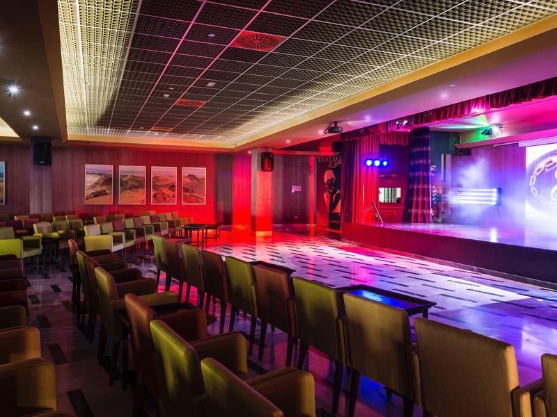 Interno di un bar elegante con palco e pista da ballo, illuminato da luci colorate.