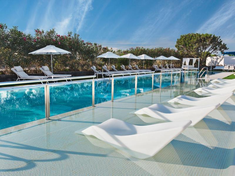 Area piscina moderna con lettini in acqua e ombrelloni sotto un cielo azzurro.