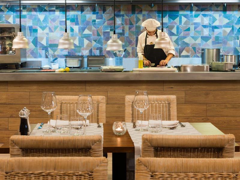 Area ristorante con tavoli apparecchiati e cuoco nella cucina con muro piastrellato blu.