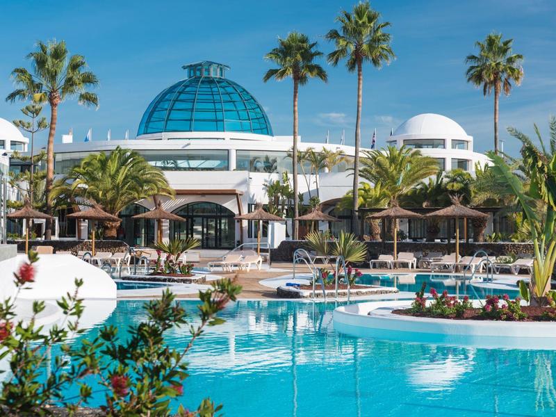 Complesso alberghiero moderno con piscina, palme e cupola blu sotto cielo limpido.