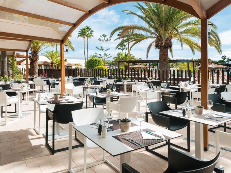 Area terrazza aperta dell'hotel con tavoli apparecchiati e vista sulle palme.
