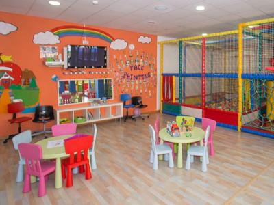 Bunter Kinderraum mit Tischen, Stühlen, Klettergerüst und Spielzeug vor orangefarbener Wand.