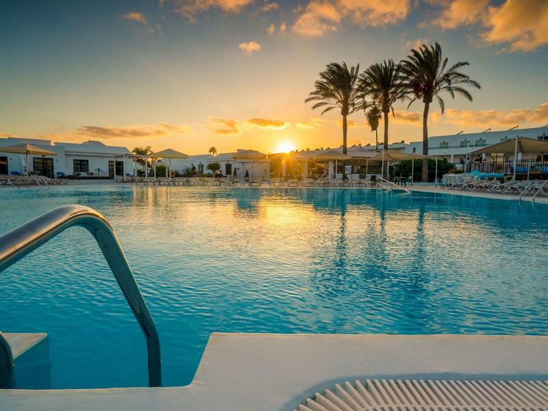 Sonnenuntergang über einem großen Pool mit Palmen und weißen Gebäuden im Hintergrund.