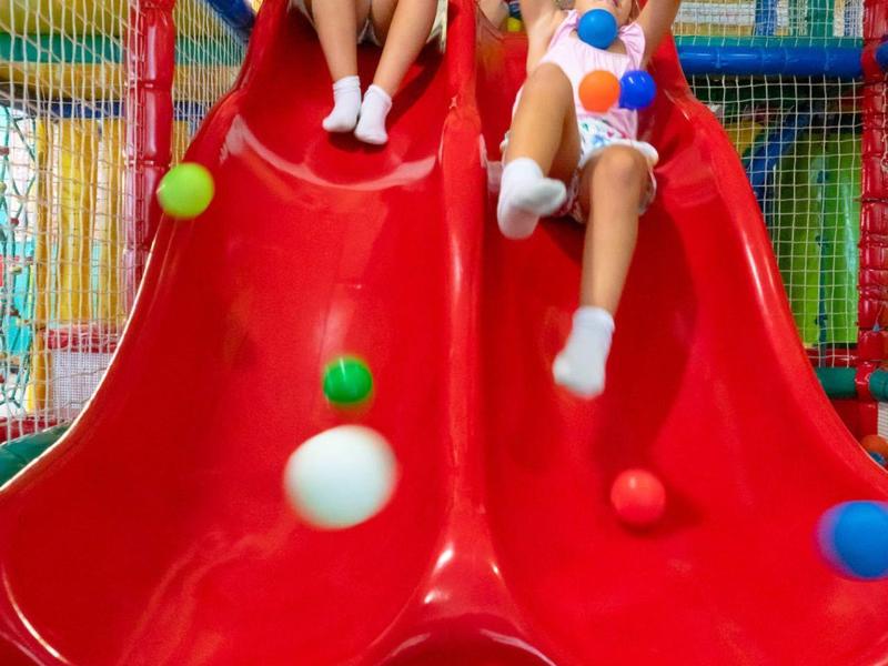 Zwei Kinder rutschen gemeinsam eine rote Rutsche hinunter in einem bunten Spielplatz mit vielen Bällen.