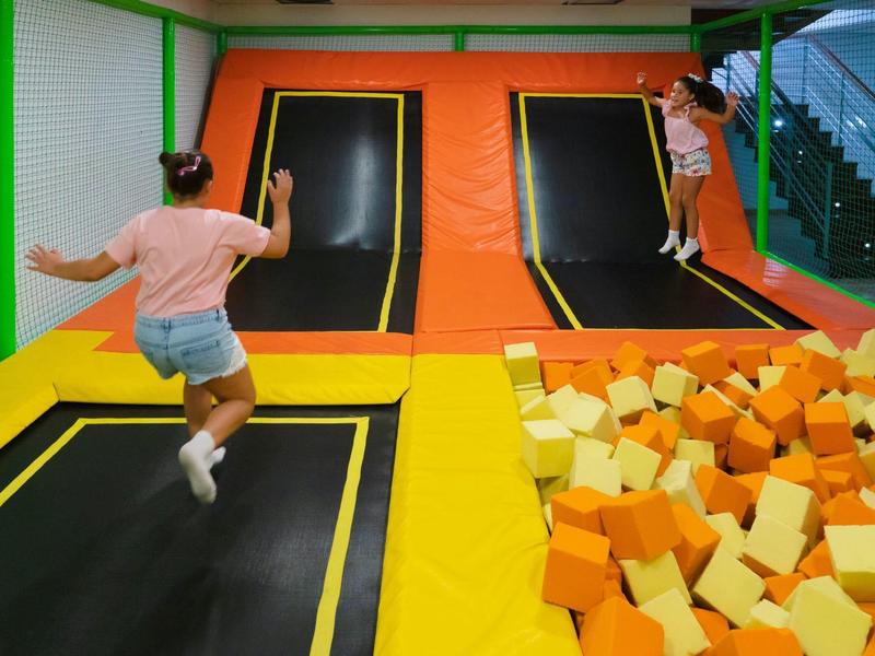 Zwei Kinder springen auf Trampolinen neben einem großen Becken mit gelben und orangefarbenen Schaumstoffwürfeln.