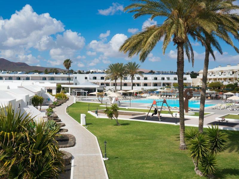 Moderner, weißer Hotelkomplex mit Pool, Palmen und grünen Rasenflächen unter blauem Himmel.