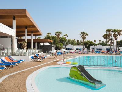 Hotel pool with water slide, sun loungers, and palm trees under clear sky.