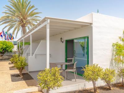 Bungalow bianco moderno con terrazza, piante verdi e palme sotto un cielo sereno.