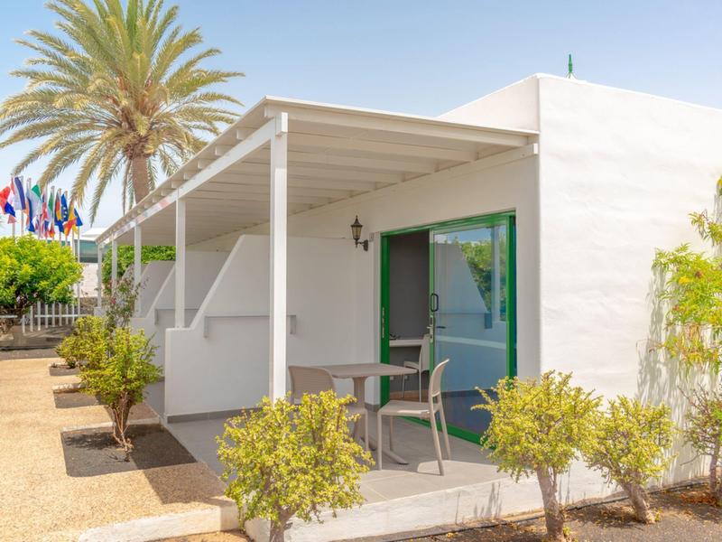 Bungalow bianco moderno con terrazza, piante verdi e palme sotto un cielo sereno.