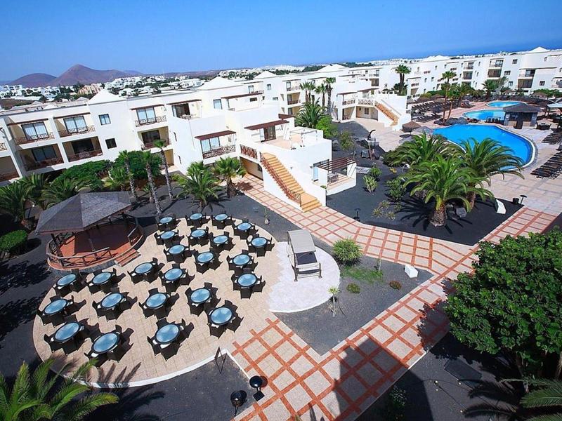 Große Hotelanlage mit Pool, Sonnenliegen, Tischen im Freien und weißen Gebäuden unter blauem Himmel.