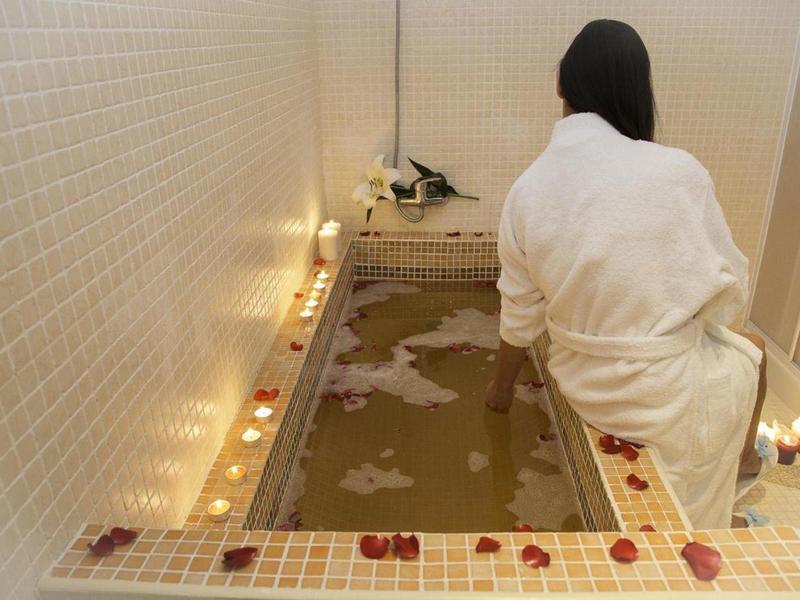 Frau in weißem Bademantel sitzt neben mit Kerzen und Rosenblättern dekoriertem Bad.