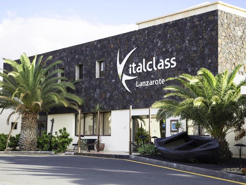 Gebäude mit dunkler Steinfassade, "italclass Lanzarote" Schriftzug, Palmen und Straße im Vordergrund.