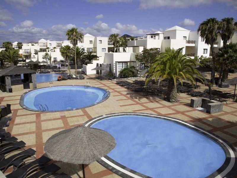 Freizeitbereich mit zwei blauen Pools, weißen Gebäuden, Palmen und Sonnenliegen unter blauem Himmel.