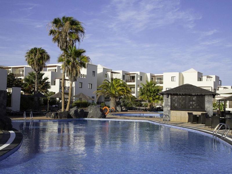 Großer blauer Pool vor weißen Gebäuden mit Palmen und blauem Himmel.