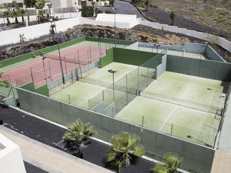 Vier Tennisplätze in einem umzaunten Bereich, zwei rot und zwei grün, umgeben von Palmen und Gebäuden.