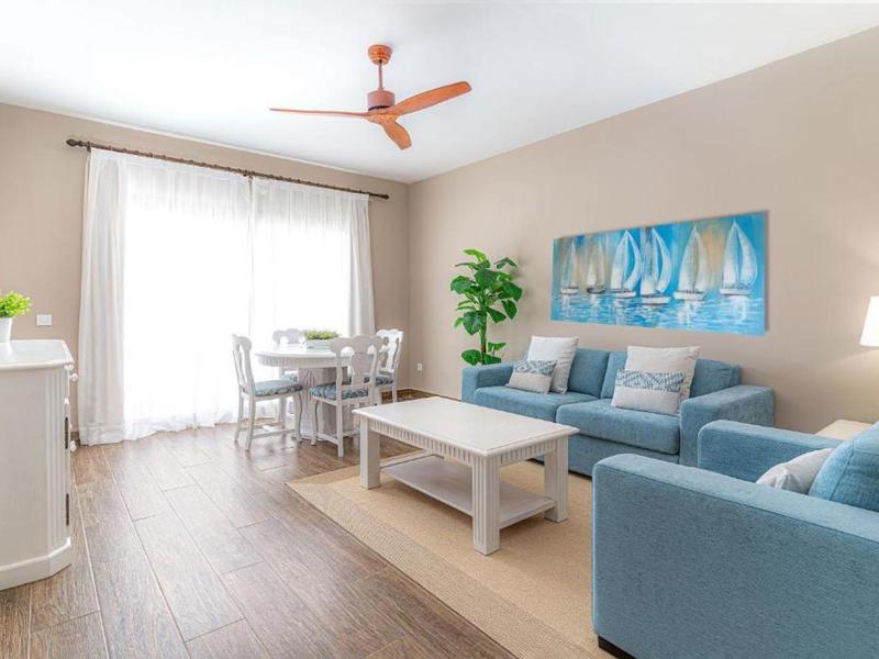 Helles Wohnzimmer mit blauen Sofas, weißem Couchtisch, Esstisch, Holzfußboden und großer Fensterfront.