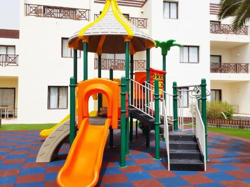 Bunter Kinderspielplatz mit Rutsche, Klettergerüst und buntem Bodenbelag vor Wohngebäude.