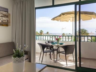 Balcone con sedie e tavolo con vista sulla piscina e palme, interno con divano e pianta.