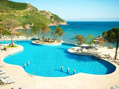 Großer, geschwungener Pool mit blauem Wasser, Liegestühlen und Blick auf das Meer und grüne Hügel.