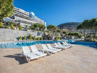 Leere weiße Liegestühle entlang eines Pools mit Hotelgebäude und Bergen im Hintergrund.