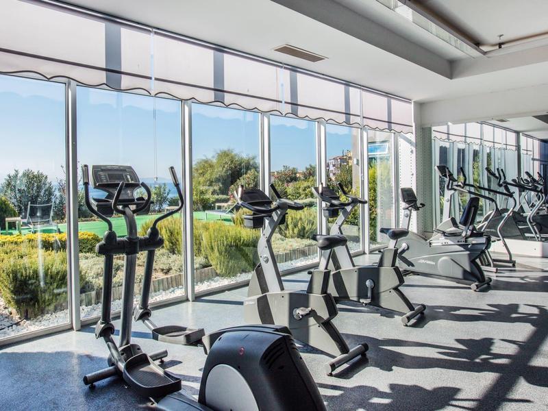 Fitnessruimte met moderne apparatuur en grote ramen met uitzicht op een groene buitenruimte.