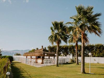 Grüne Wiese mit Palmen und einem überdachten Sitzbereich nahe einem Zaun und Bergen im Hintergrund.