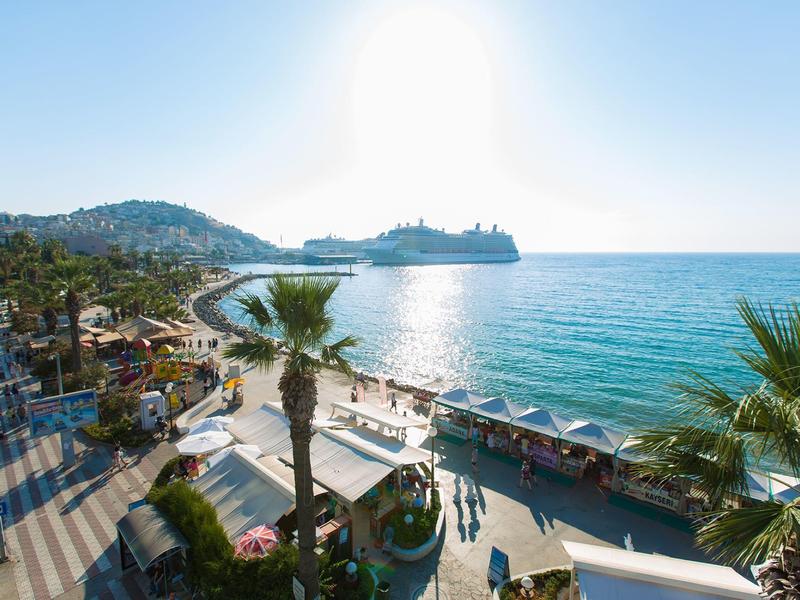 Nasłoneczniony odcinek plaży z promenadą, palmami i widokiem na morze oraz wzgórza w tle.