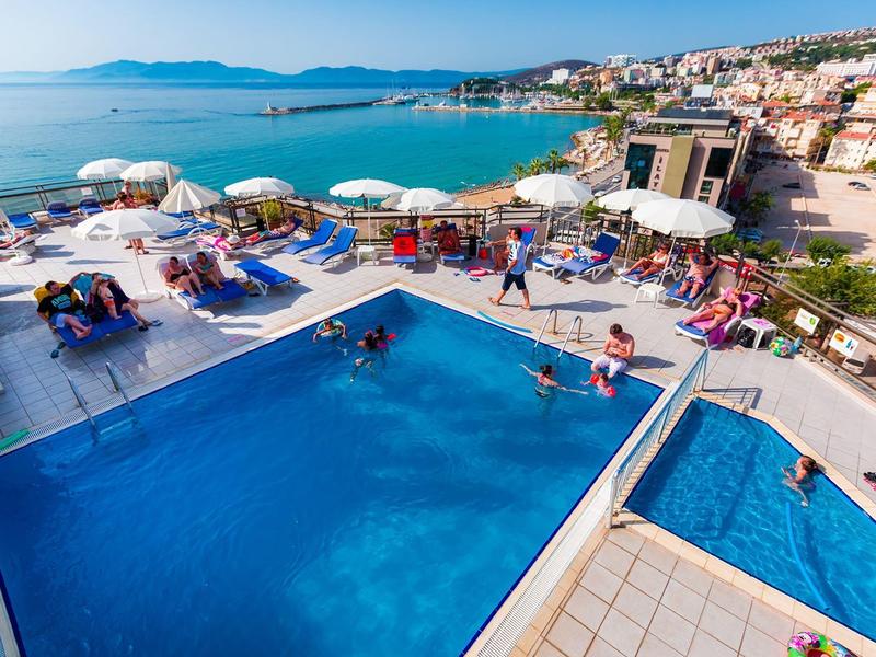 Basen na zewnątrz z leżakami i parasolami, widok na morze i miasto w tle.