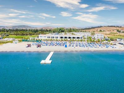 Hotel bianco sulla spiaggia con lettini, ombrelloni e un molo che si estende nel mare blu chiaro.