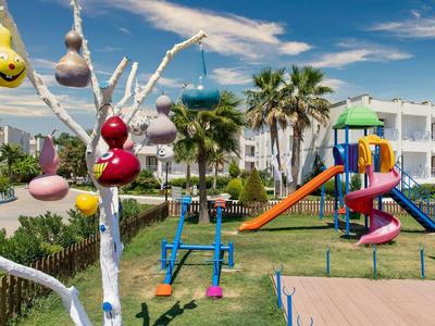 Area giochi colorata per bambini con scivoli e altalene davanti a un hotel con palme.