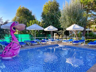 Piscina con acqua blu, lettini, ombrelloni bianchi e un gonfiabile fenicottero rosa sotto gli alberi.