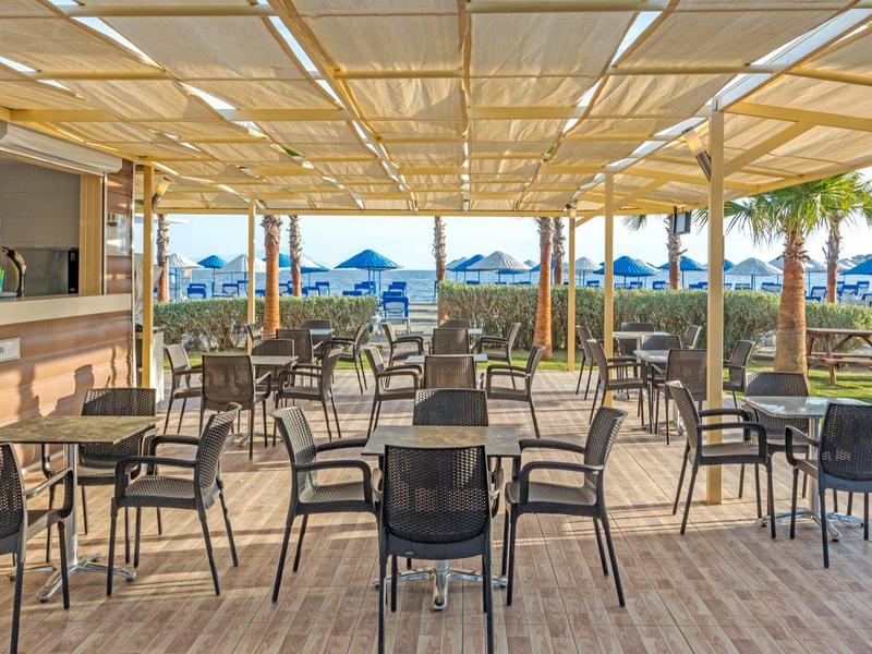 Terrazza coperta con sedie e tavoli con vista sulla spiaggia e sul mare, palme sullo sfondo.