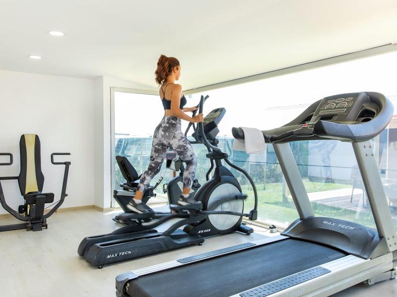 Donna si allena su un attrezzo ellittico in una palestra luminosa con grandi finestre con vista esterna.