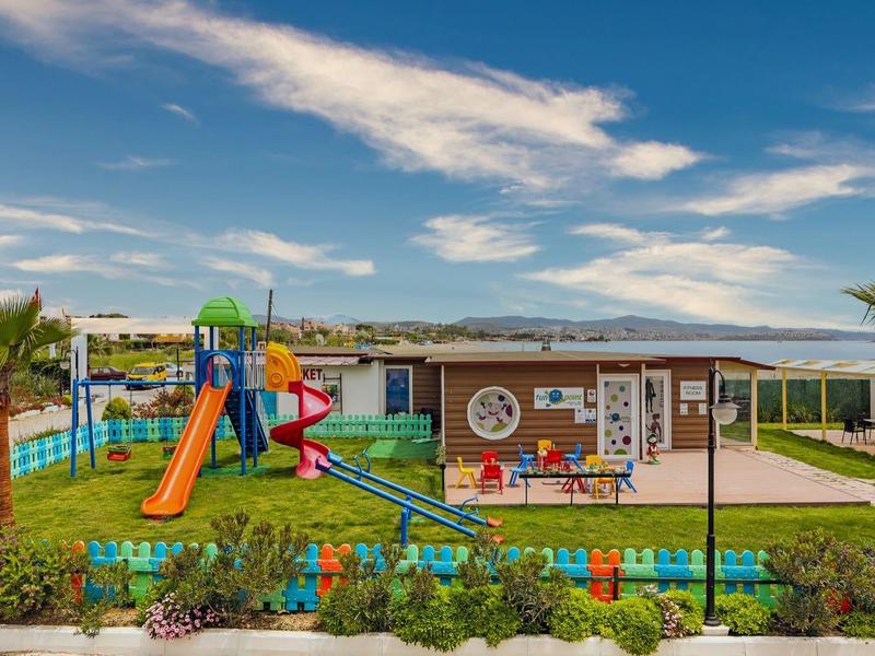 Parco giochi colorato per bambini davanti a un hotel con palme e vista sulle montagne.
