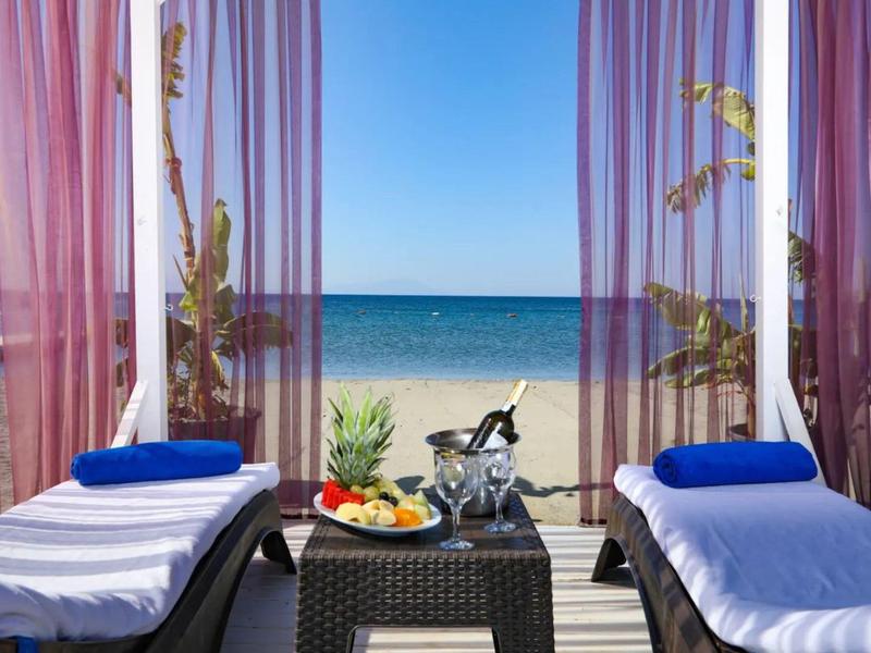 Due lettini con asciugamani blu di fronte a una spiaggia con vista mare e un piatto di frutta.