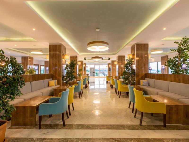 Ampia sala da pranzo con sedie colorate, tavoli, piante, ben illuminata e accogliente.