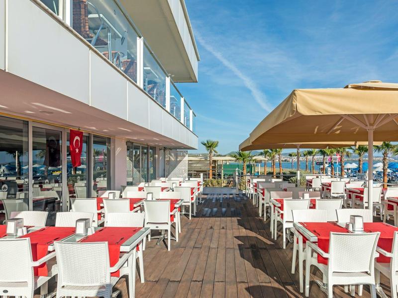 Terrazza di un hotel con sedie bianche, tovaglie rosse e vista piscina sotto cielo sereno.