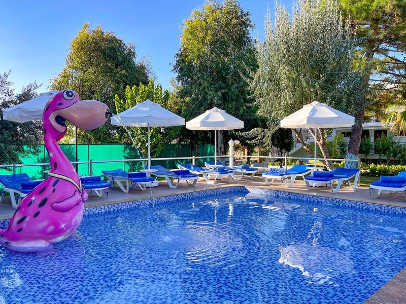 Piscina con acqua blu, lettini, ombrelloni bianchi e un gonfiabile fenicottero rosa sotto gli alberi.