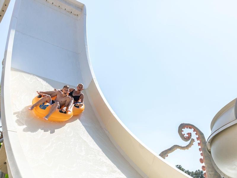 Deux personnes glissent sur un toboggan aquatique blanc avec un ciel bleu clair en arrière-plan.