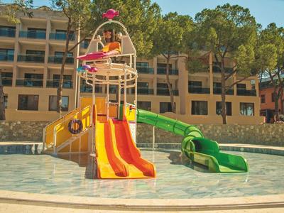 Kinderwasserrutsche mit gelben und grünen Rutschen im flachen Pool vor Hotelgebäude und Bäumen.