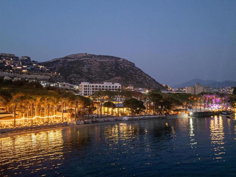 Wieczorna nadmorska promenada z oświetlonymi budynkami i wzgórzem w tle.